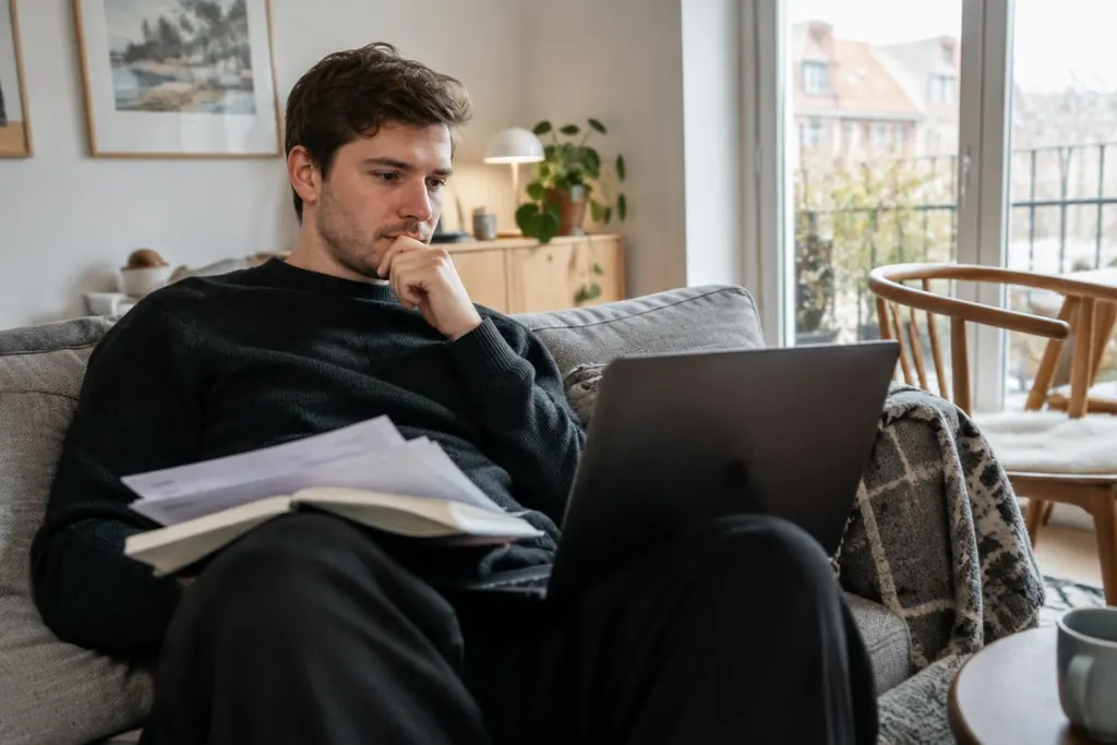 En ung mand overvejer sin økonomi med computer og regninger i sofaen.